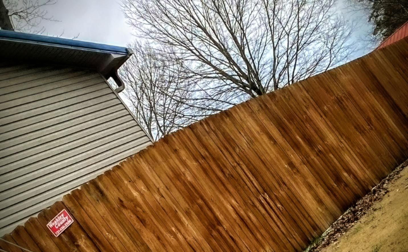 Tall stained wood privacy fence running beside a house, with a red Weakley County Fence sign on the first panel.